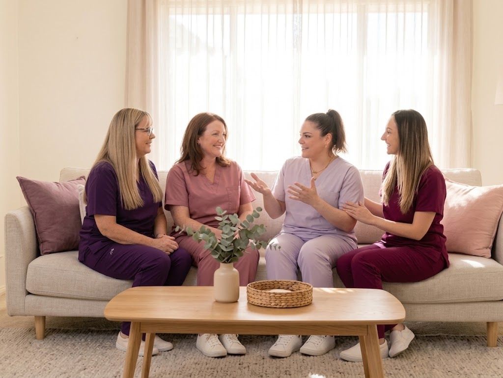 The four EverNurture midwives sitting together on a sofa in their scrubs, talking and laughing