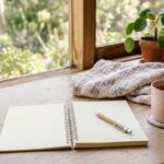An open notepad with a pen, a mug of tea and a folded muslin wrap on a timber bench beside a sunlit window